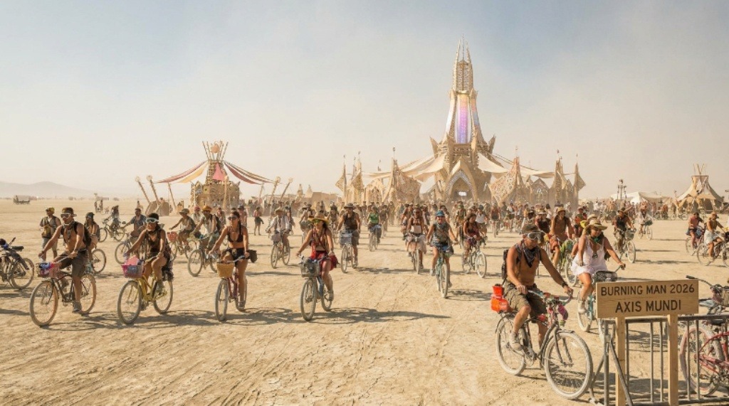 Participants on bicycles crossing the open playa at Burning Man 2026 Axis Mundi with art installations in the background
