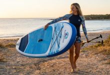 Woman carrying a lightweight inflatable paddle board backpack on beach.